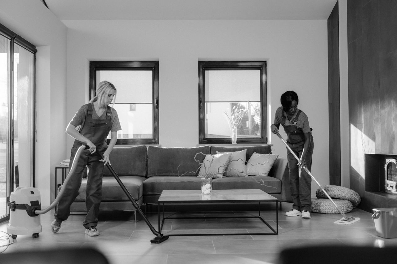 Two women in work clothes cleaning a modern living room with a vacuum cleaner and mop.